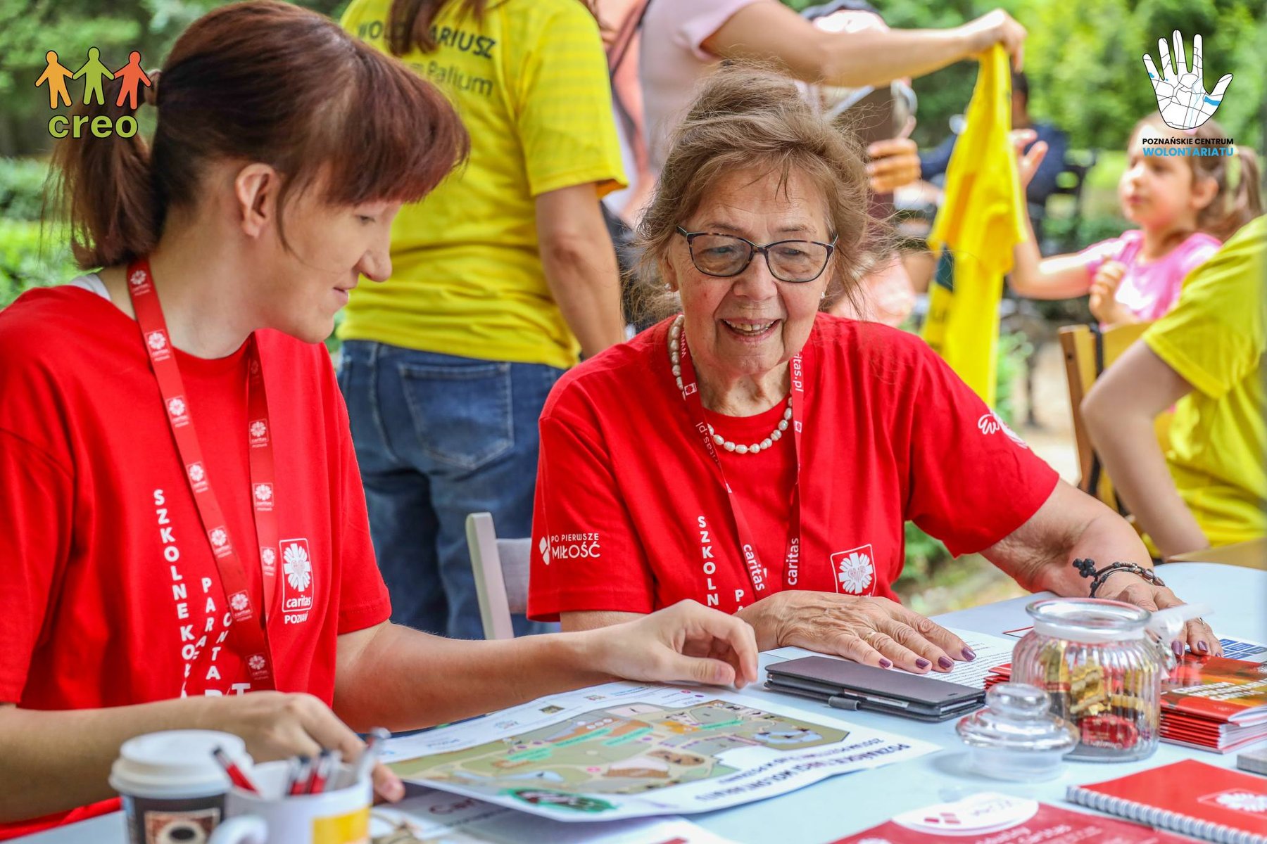 W parku Wilsona czekać będą trzy strefy: wystawiennicza, warsztatów i spotkań oraz doświadczeń i animacji/ fot. CREO Na zdjęciu dwie wolontariuszki w czerwonych koszlkach siedzące przy stoliku, w tle dzieci