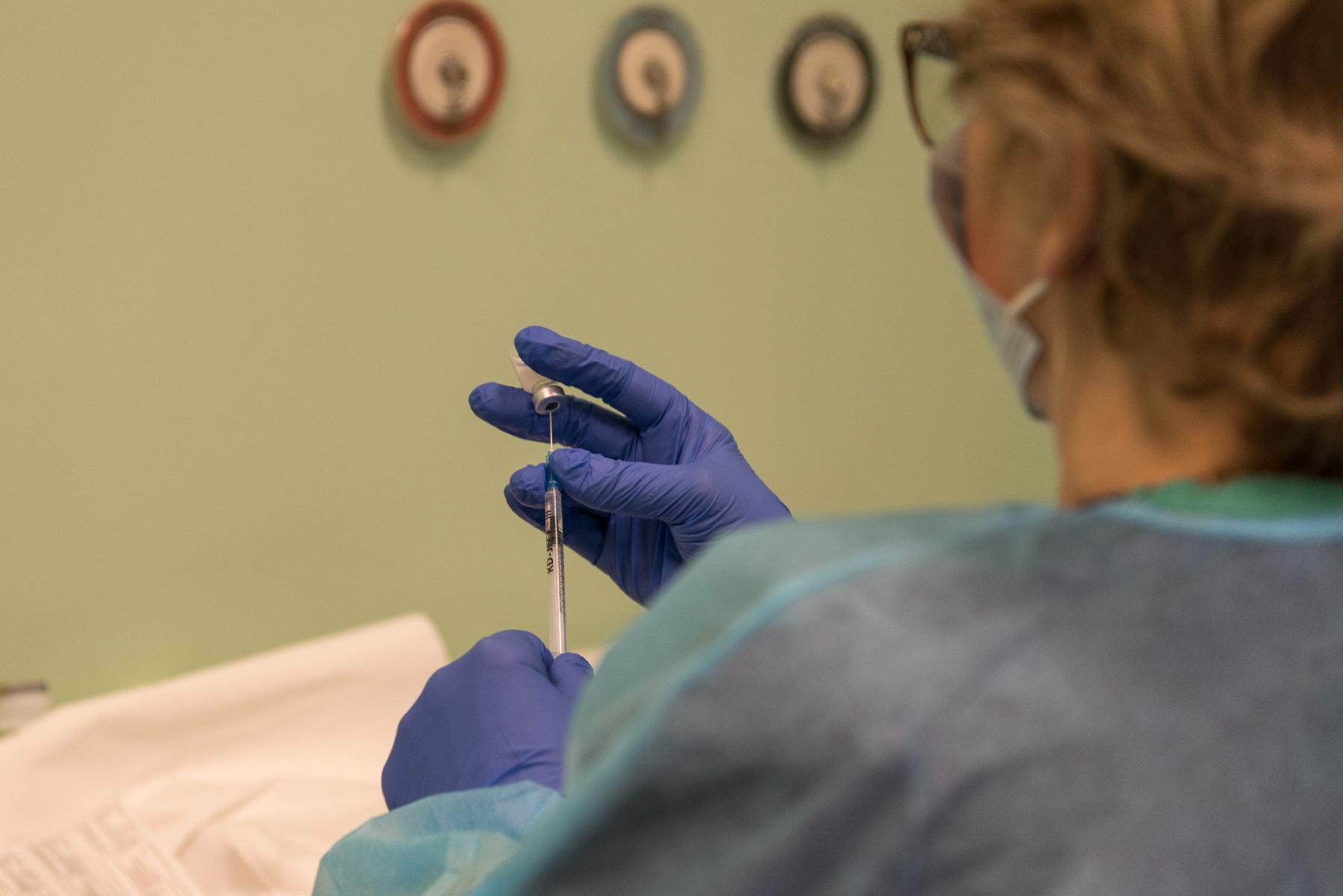 Stojąca tyłem kobieta w medycznym fartuchu, jedną dłonią (w rękawiczce) trzyma szczepionkę, drugą - nabiera ją strzykawką - grafika artykułu