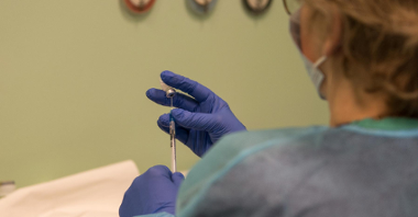Stojąca tyłem kobieta w medycznym fartuchu, jedną dłonią (w rękawiczce) trzyma szczepionkę, drugą - nabiera ją strzykawką