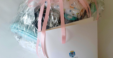 Na zdjęciu prezenty opakowane w przezroczystą folię, przewiązane różową wstążką, przed nimi stoi teczka z listem gratulacyjnym