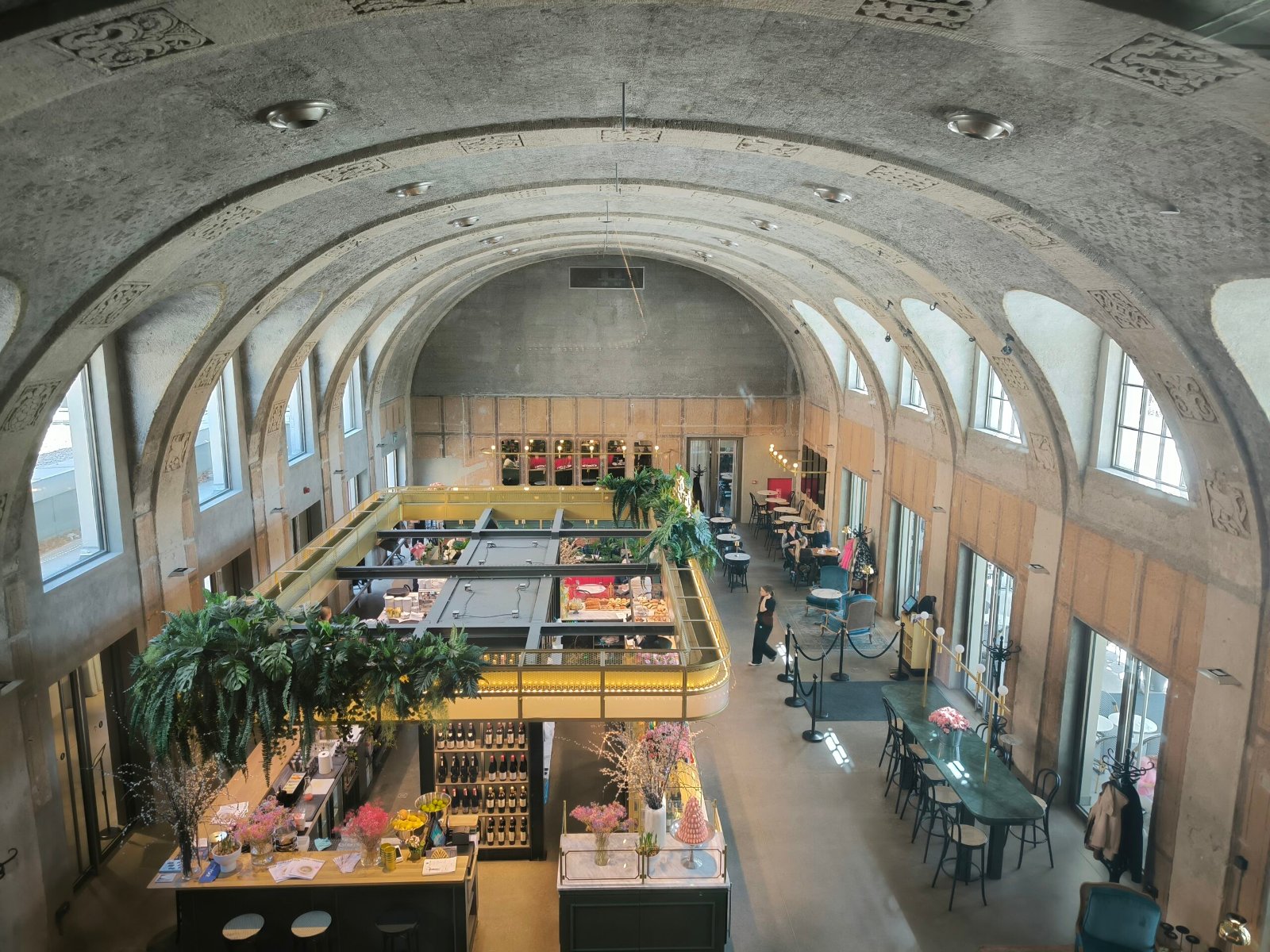 Znamy wyniki konkursu "Zabytek Zadbany"/ fot. NID/D. Mikulski Wnętrze modernistycznego budynku z wysokim, łukowatym sufitem i rzędami okien, mieszczącego restaurację lub kawiarnię. W centralnej części widać otwartą kuchnię lub bar z wiszącymi roślinami i oświetleniem. Rozstawione są stoliki i krzesła, a w tle widać wejście i dalszą część lokalu. Architektura wnętrza sugeruje historyczny budynek, przekształcony na cele gastronomiczne.