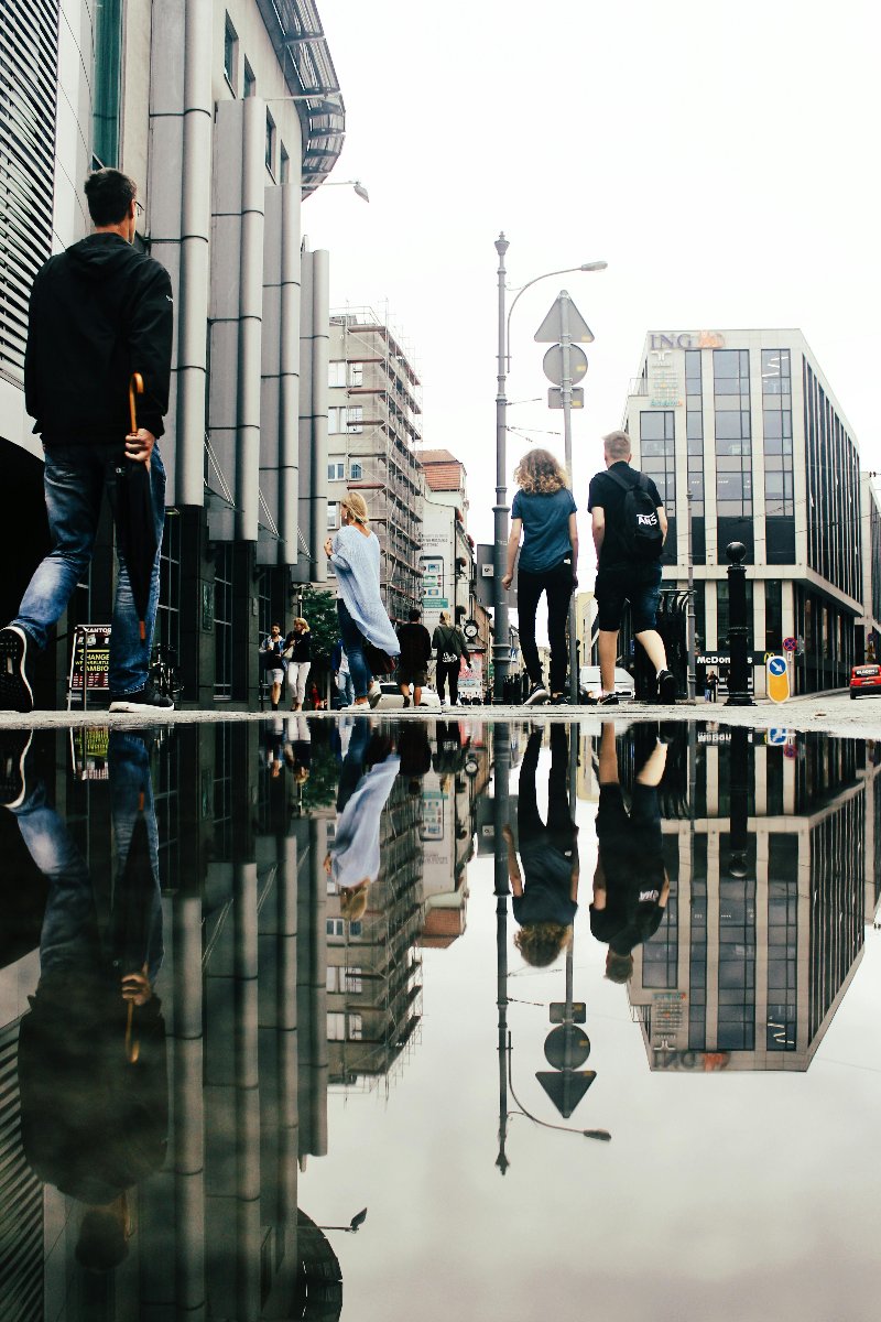 Centrum miasta, okolice Kupca Poznańskiego i ulicy Podgórnej. Zdjęcie zostało zrobione od dołu z poziomu chodnika. Z lewej strony widać część galerii Kupiec Poznański. Na wprost widać ulicę Półwiejską. Z lewej strony idzie mężczyzna z parasolem. W oddali widać spacerujących przechodniów. Na dole zdjęcia została uchwycona kałuża, w której odbija się opisany obraz.