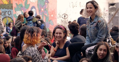 Grupa ludzi otoczonych ścianami z kolorowym grafitti. Z przodu grupa roześmianych dziewczyn.