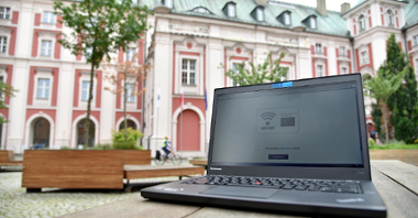 Zdjęcie przedstawia dziedziniec Urzędu Miasta Poznania. Na pierwszym planie widać komputer (laptop), stojący na ławce - na ekranie wyświetlona jest strona umożliwiająca połączenie z siecią, w tle widać budynek urzędu