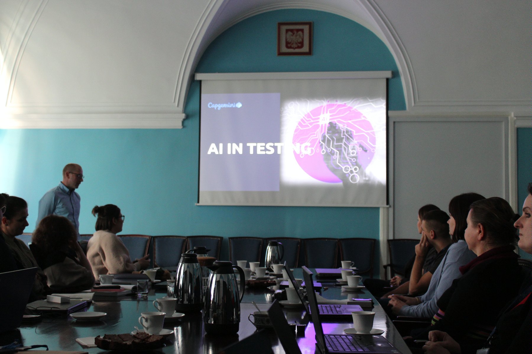 Na obrazie grupa ludzi siedzących przy stole patrzy na prezentację wyświetlaną na ekranie oraz słucha prowadzącego szkolenie - grafika artykułu