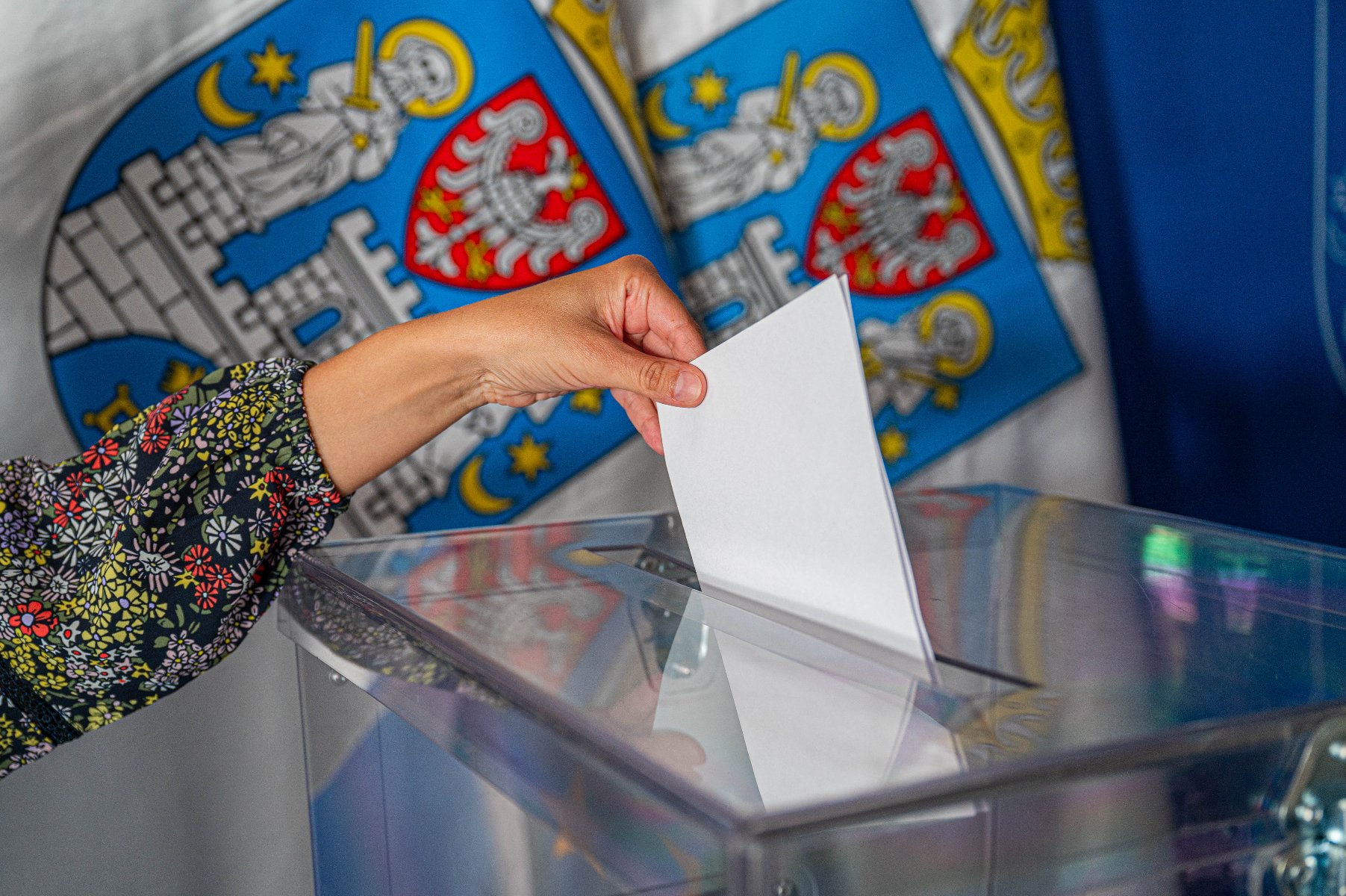 Zdjęcie przedstawia rękę wrzucającą kartę do głosowania do urny wyborczej. W tle flagi z herbem Miasta Poznania. - grafika artykułu