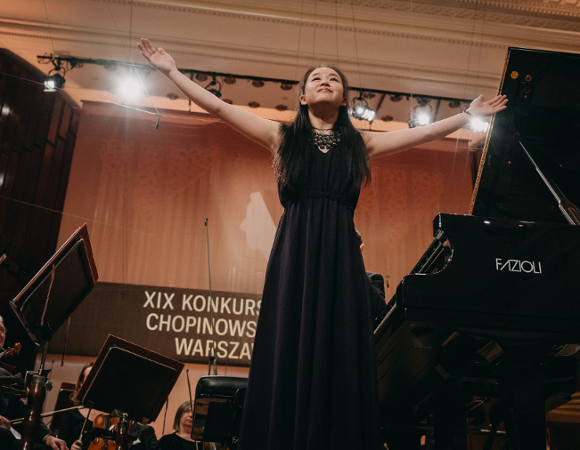 Tianyao Lyu, studiująca w Poznaniu Chinka, zajęła 4. miejsce podczas XIX Międzynarodowego Konkursu Pianistycznego im. Fryderyka Chopina fot. chopincompetition.pl/Wojciech Grzedziński młoda pianistka w czarnej sukni stoi przy fortepianie na scenie z uniesionymi rękami w geście podziękowania