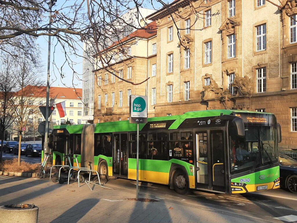 . przed budynkiem stoi zielono-żółty autobus miejski