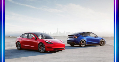 Dwa samochody marki Tesla. Po prawej niebieski, po prawej czerwony. Samochody są oświetlone z prawej strony. W tle panomara miasta.
