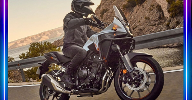 Mężczyzna w stroju motocyklisty jedzie na czarnym motocyklu. W tle góry i jezioro.