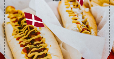 Dwa hot dogi w papierkach. Każdy z nich ma długą parówkę, jest polany musztardą i keczupem. W parówki wbite są flagi Danii.