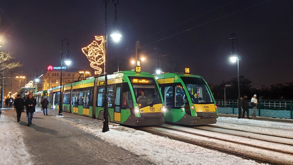 At a snowy tram stop at night, two modern green-and-yellow trams are standing side by side on the tracks. Streetlights illuminate the area, and one of the lampposts is decorated with a glowing Christmas angel light. Snow covers the ground and the rails. A few pedestrians in winter clothing are visible nearby. The scene feels calm, wintry, and festive.