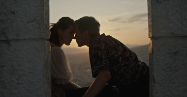 Picture from the movie - a teenage girl and boy touching with their foreheads, between two stone walls at dusk.