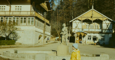 Photo of a girl sitting on a concrete parapet in front of a sculpture. Two buildings in a background.