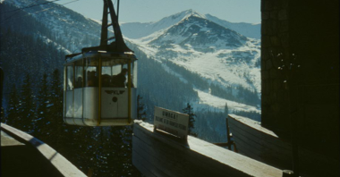 Picture of a cable car reaching a station. Snow-peaked mountains as a background.
