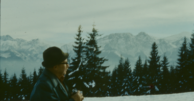 Picture of the photographer, Władysław Rut, in a profile, holding a camera in his hands. Green spruce trees and snow-peaked mountains as a background.