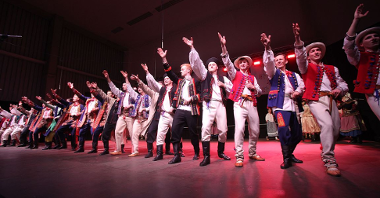 Photo of a folk group in traditional folk costumes performing on stage - dozen or so men standing in a row with their right hands up.