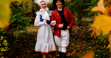 Photo of a woman and a man dressed in traditional folk costumes. Trees and colourful leaves as a background.