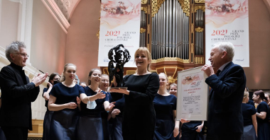 Picture of the winners - the choir's woman conductor holding a statuette and two men - one of them holding a diploma and the other clapping his hands. Behind the conductor girls members of the choir. Pipe organ in the background.