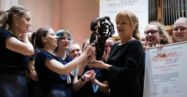 Photo of a woman conductor holding a statuette, girl members of the choir around her, one of them holding a diploma.