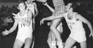 Black and white photo of five men playing basketball. One of them in a jump.