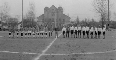 Black and white photo of two teams standing in a row on a football field. Building and trees in the background.