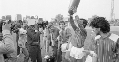 Black and white photo of football players, one of them is lifting a trophy. Opposite him several men, including one with a camera.