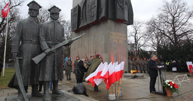 Photo of the monument to the Greater Poland Insurgents. A man speaking in front of the monument and white and red Polish flags, soldiers around the monument.