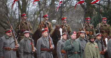 Photo of standing soldiers, behind them soldiers on horses. Trees as a background.