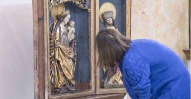 A young woman visiting the exhibition leans over the exhibit - a wooden altar closed in a display case, on which there is a figure of the Virgin Mary with Jesus. The figure is richly decorated and full of gold.