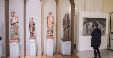 Four wooden figures of different sizes placed next to each other. Three of them depict images of Mary. Next to them there is a large graphic painting depicting a mutilated face.