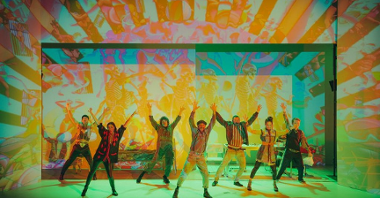 Photo shows a frame from the performance - actors with their hands up, against a backdrop of coloured lights