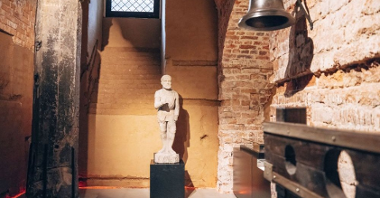A statue of a knight, next to it there was a pedestal on which a sculpture stood. On the right, a medium-sized bell hangs on the brick wall.