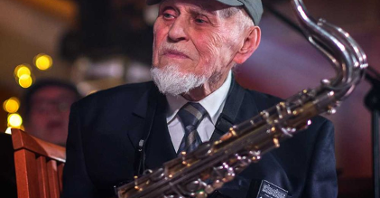 Photo of the elderly man in a peaked cap, holding a saxophone. The man is looking aside.