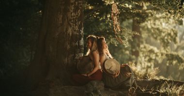 Two girls in dresses are sitting under a large tree, leaning against each other, each holding an old-fashioned drum in her arms. One of them is illuminated by the sun.