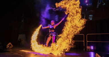 A woman in a leather outfit stands on the stage holding two torches in her hands, from which a huge fire bursts.
