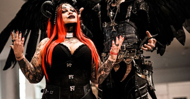 A couple in gothic costumes stand presenting their outfits. The woman has long, red hair and black horns on her head, the man has many earrings, big, black wings and diabolic makeup.