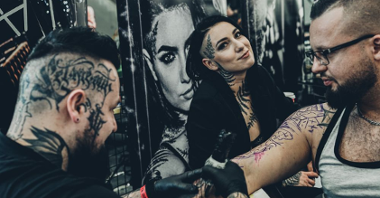 Two men and one woman, one of men works as a tattoo artist. The woman smiles to a camera, the men are busy tattooing.