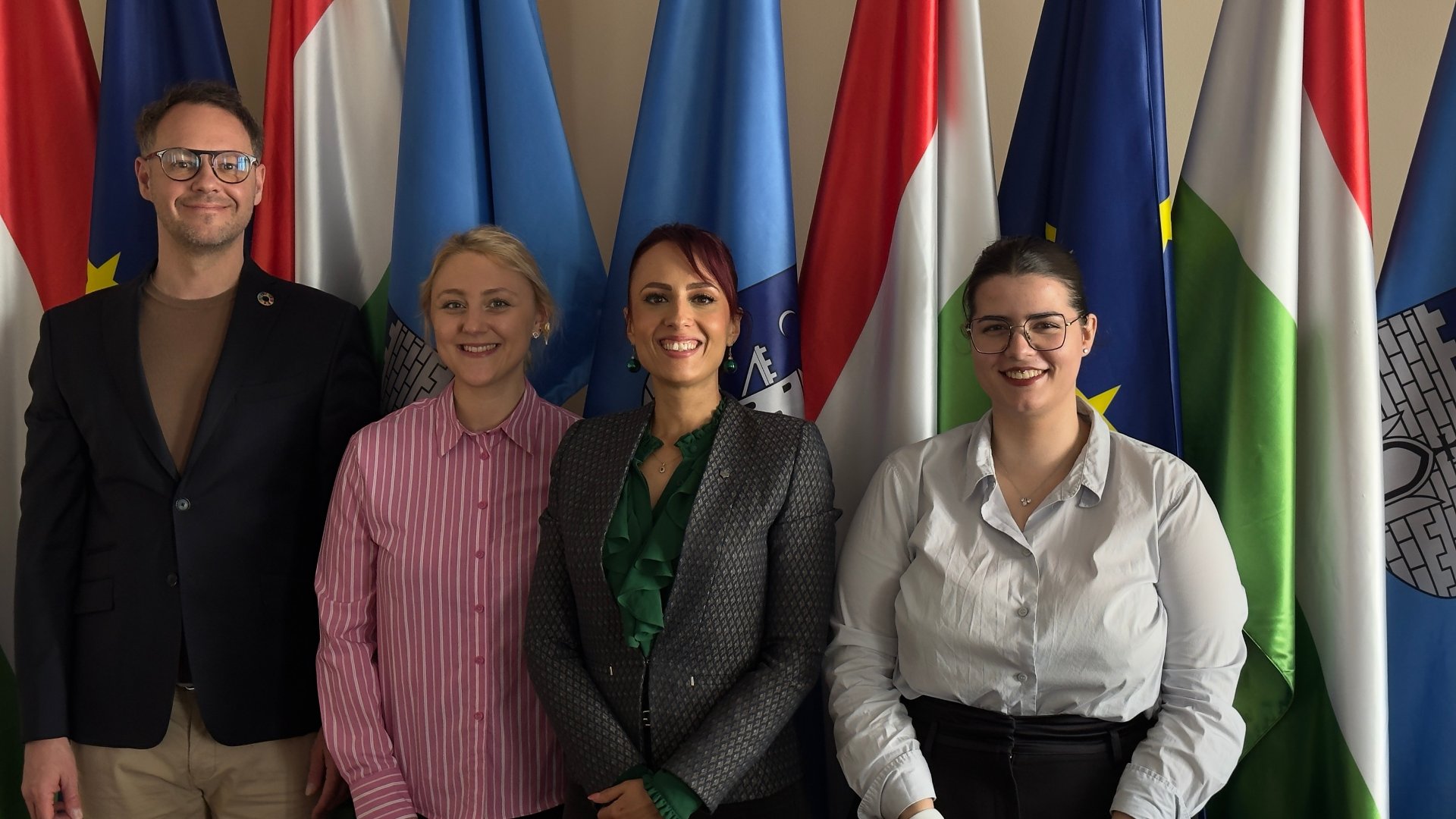 The photo shows three smiling women and a man against a background of several flags - grafika artykułu