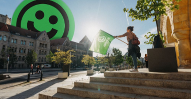 A woman with a green flag in her hands stands on the steps in front of the building. In the background there is a street and a historic building with a smiling face emoticon sticking out from behind it.
