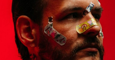 A photo of a man's face with colorful plasters glued to his nose, cheeks and forehead. Red background.