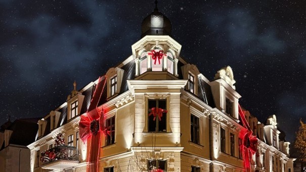 The image shows a historic building decorated for Christmas, illuminated with lights highlighting its architectural details. Large red ribbons and bows are draped across the facade and windows, giving the appearance of a giant holiday gift. A balcony is decorated with a Christmas tree and ornaments, adding to the festive atmosphere.