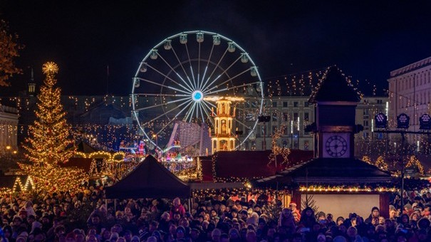 The photo shows a Christmas market at night, filled with a large crowd of people enjoying the festive atmosphere. The scene is beautifully illuminated with countless Christmas lights decorating the stalls and surrounding buildings. A giant Ferris wheel and a brightly lit Christmas tree dominate the background, adding to the magical holiday ambiance. Small wooden market huts and a clock tower are also visible, creating a traditional festive setting. The market is lively, cheerful, and full of holiday spirit.