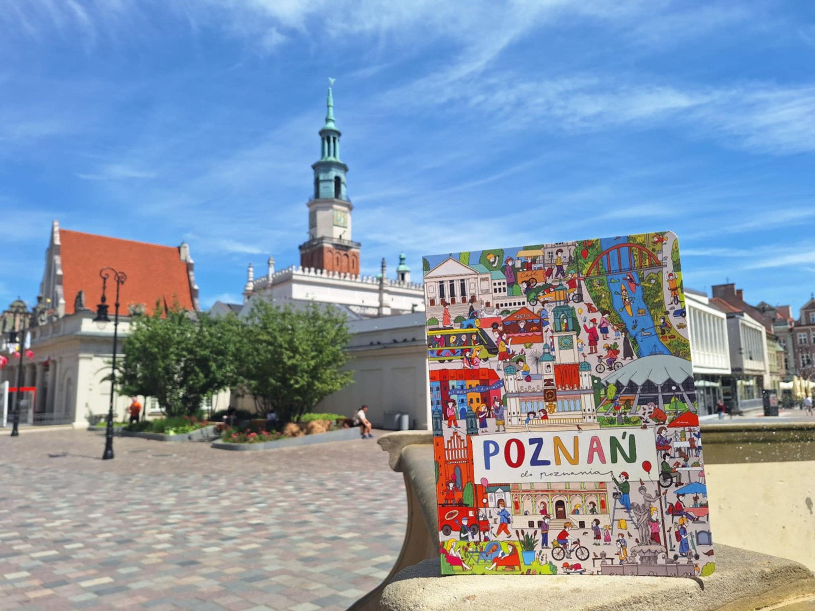 "Poznań do poznania" nowość wydawnicza dla najmłodszych Książka stoi na murku stanowiącym fragment fontanny. W tle zabytkowy stary rynek z okazałą wieżą ratuszową