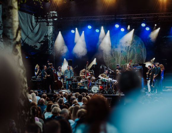W przyszłym tygodniu rzusza kolejny festiwal muzyczny. Od 15 do 18 czerwca wybrzeża Jeziora Strzeszyńskiego rozgrzeje Enter Enea Festival. Na pierwszym planie głowy słuchaczy, którzy siedzą pod sceną i oglądają koncert. Na scenie zespół składający się z wielu muzyków. Scena jest oświetlona bialymi i niebieskimi światłami.
