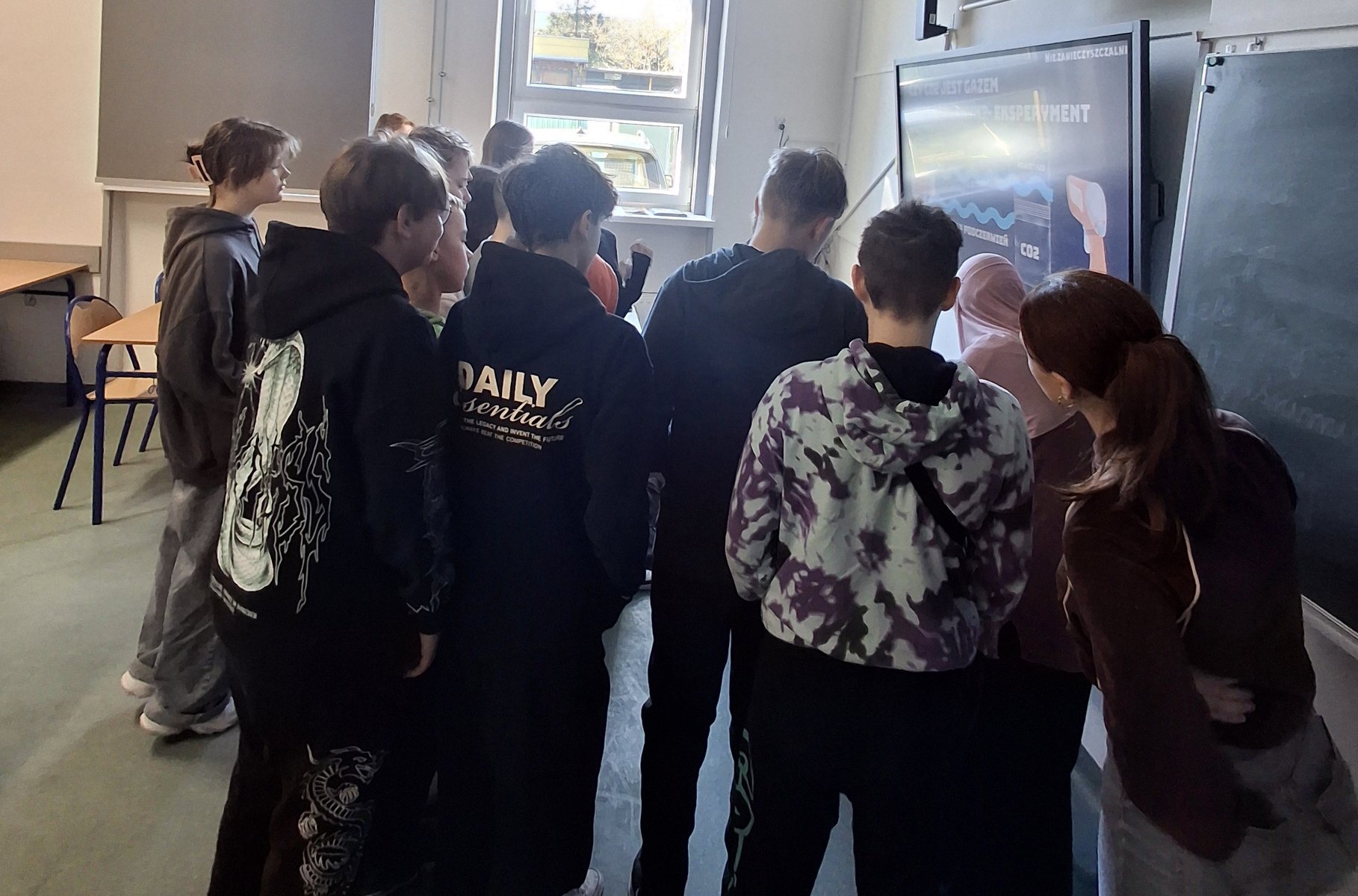 Prejekt Nie.zanieczyszczalni poznańskie licealistki realizowały w szkołach podstawowych i ponadpodstawowych Na zdjęciu widać grupę młodzieży stojącą przy szkolnej tablicy i obserwującą działania niewidoczne w kadrze