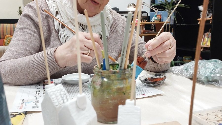 Seniorka maluje kamieniczkę przy stole.