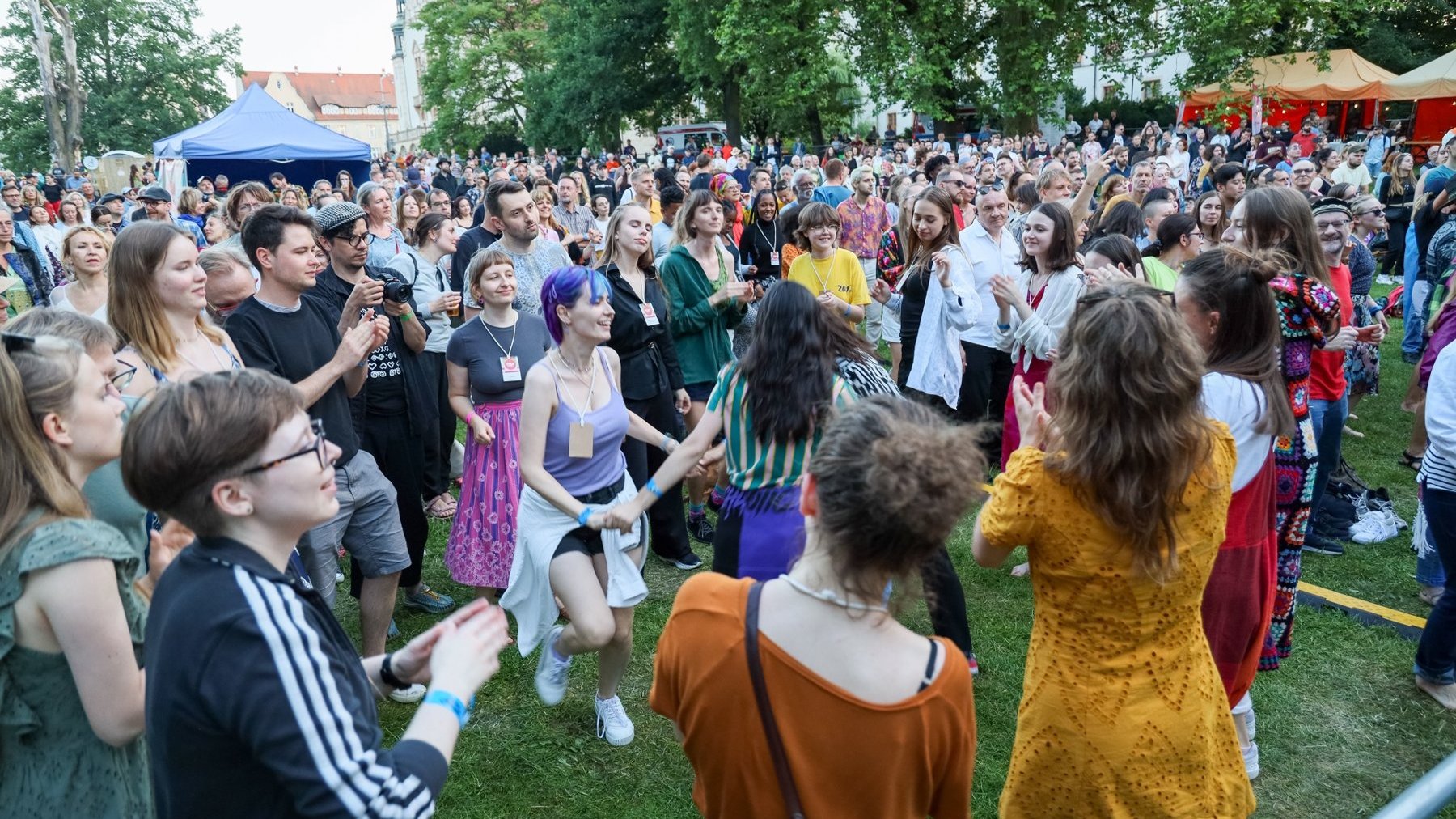 Tłum ludzi stoi w parku, na pierwszym planie widać grupę osób stojącą w kole. Klaszczą w dłonie i tańczą.