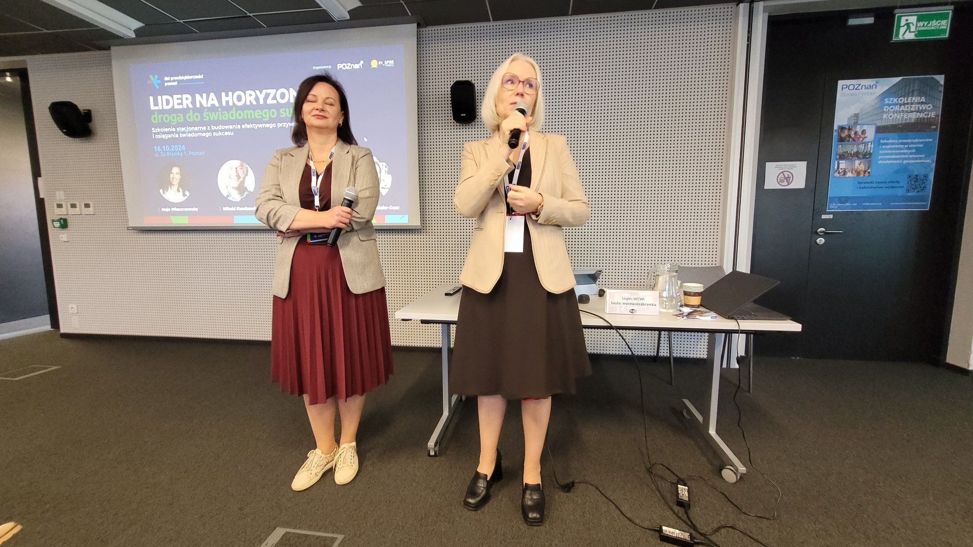 Dwie przedstawicielki fundacji In_Spire witają publiczność na konferencji "Lider na horyzoncie", jedna z nich przemawia przez mikrofon. W tle wyświetlana jest grafika promująca konferencję oraz plakat promujący Poznań Biznes Partner.