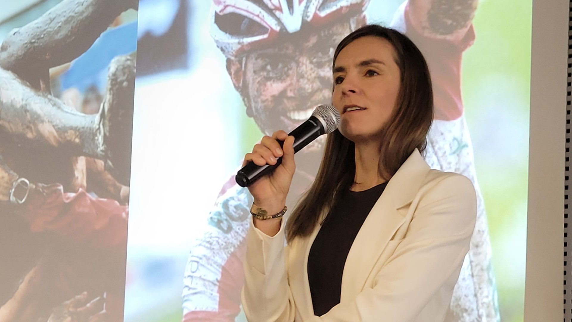 Medalistka olimpijska w kolarstwie górskim przemawia przez mikrofon podczas swojej prelekcji na konferencji "Lider na horyzoncie". W tle wyświetlane jest jej zdjęcie ze zwycięskiego wyścigu. Na zdjęciu jej twarz i ręce są pokryte błotem.